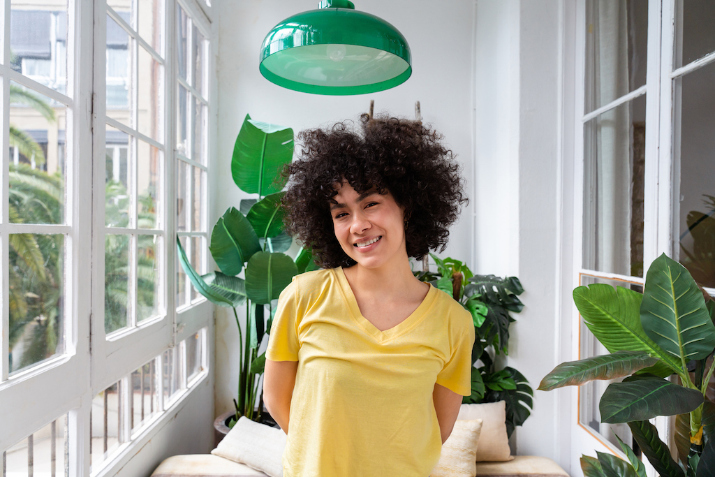 Happy woman smiling into camera with plants and green light fixture thanks to reliable residential electrician service.